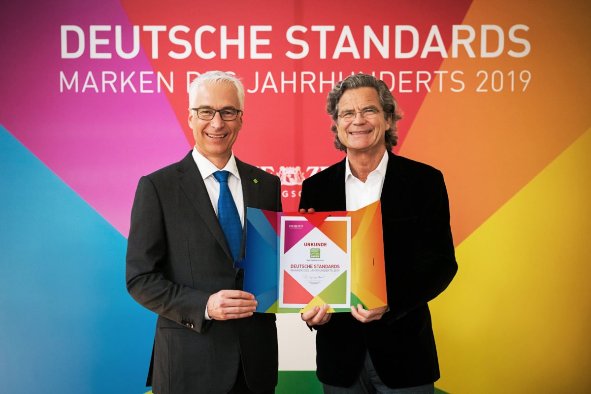Neher posiert mit Urkunde vor buntem Hintergrund mit Marke des Jahrhunderts 2019