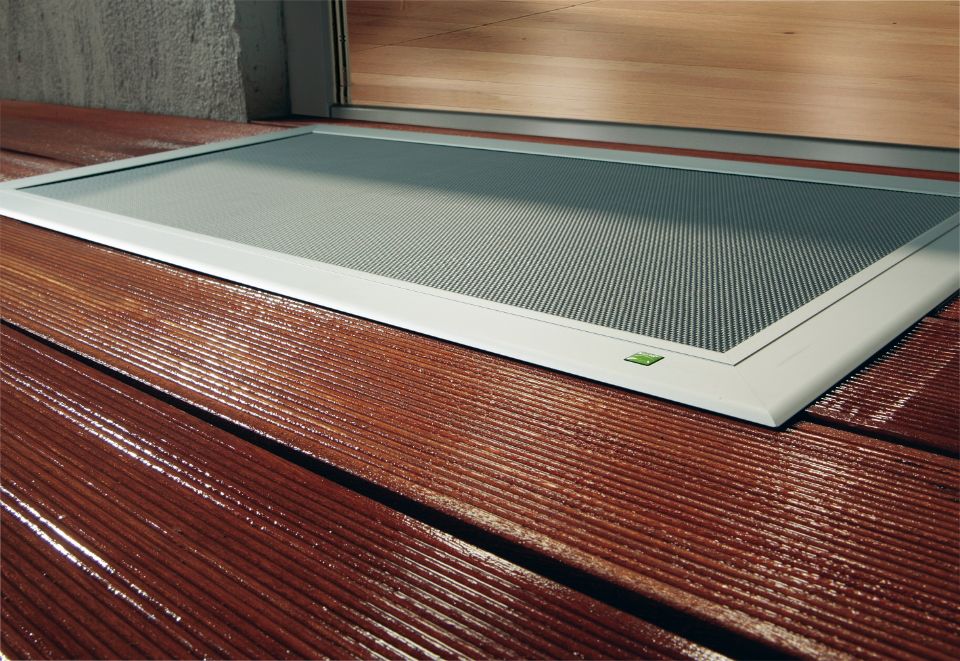 TERRESA Lichtschachtabdeckung mit elegantem Aluminiumrahmen, bündig auf einer Holzterrasse vor der Tür installiert.