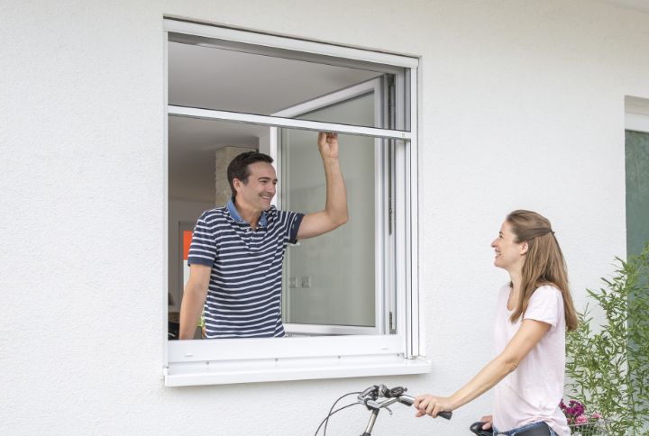 Mann bedient ein Insektenschutz‑Rollo am Fenster, während eine Frau draußen mit Fahrrad steht; leichtgängige Führungsschienen, freie Durchreiche und klare Sicht.