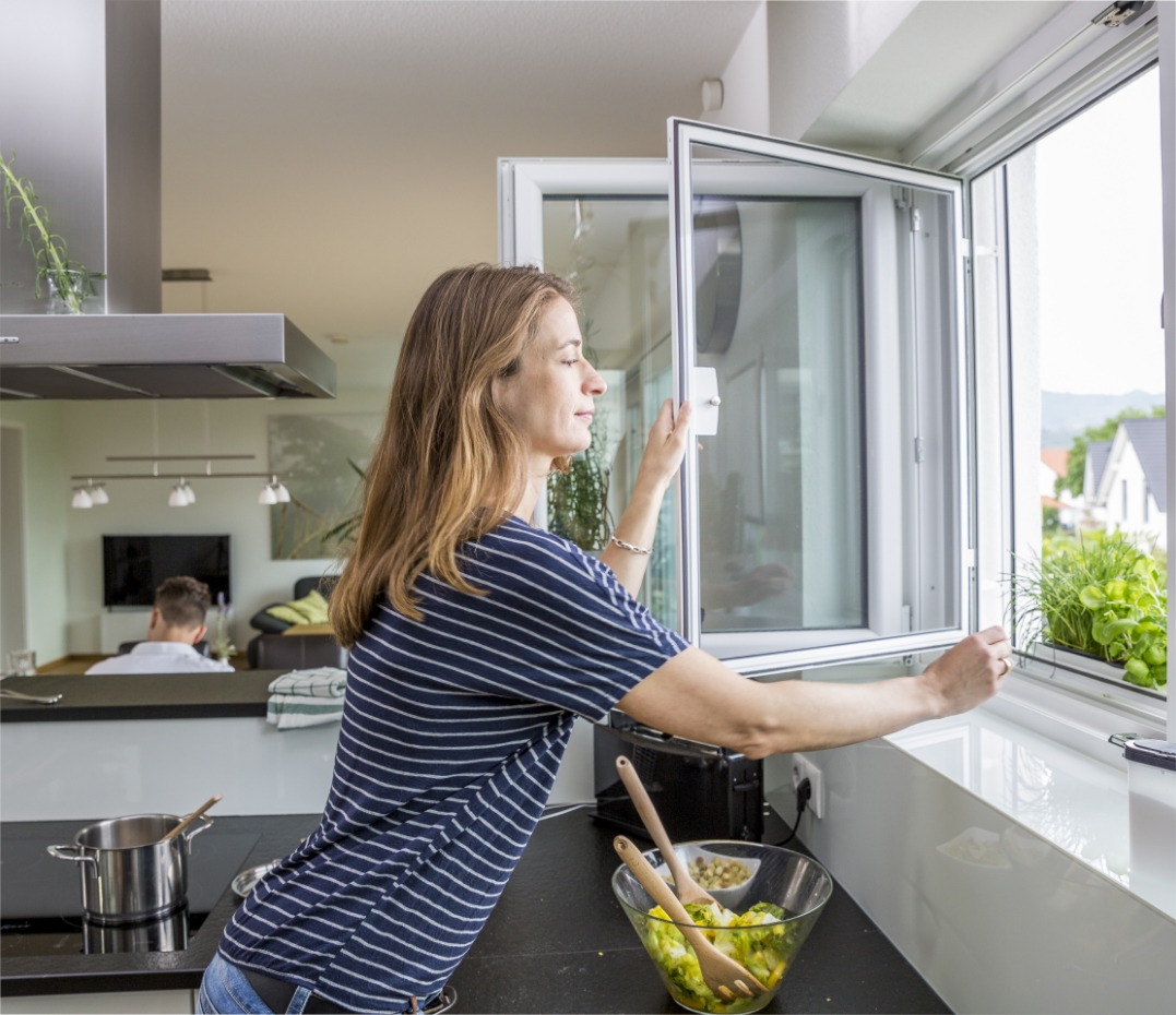 Frau steht am Fenster in einer modernen Küche und öffnet den Insektenschutz, um an Kräuter auf der Fensterbank zu kommen.