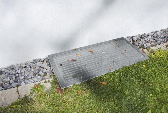 LISA Lichtschachtabdeckung aus Aluminium mit Edelstahlgewebe, flach auf dem Lichtschacht vor einer Hauswand montiert. Laub liegt auf dem Gitter.