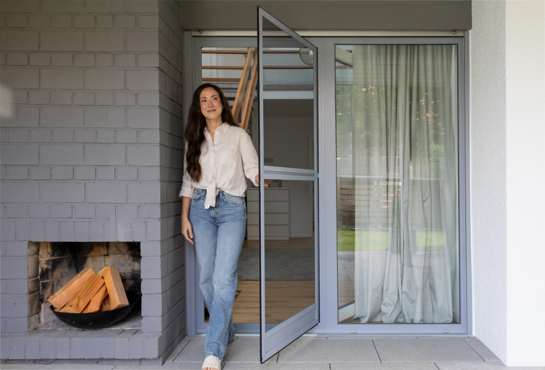 Neher Insektenschutz-Pendeltür mit Sprosse zur zusätzlichen Stabilität; Frau tritt auf die Terrasse, schlanke Aluminiumprofile und transparentes Gewebe.