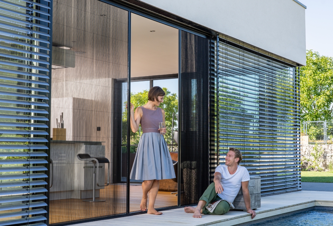 Person bedient Neher Insektenschutz Plissee an einer großen modernen Schiebetür. Eine weitere Person sitzt am Pool auf dem Boden.