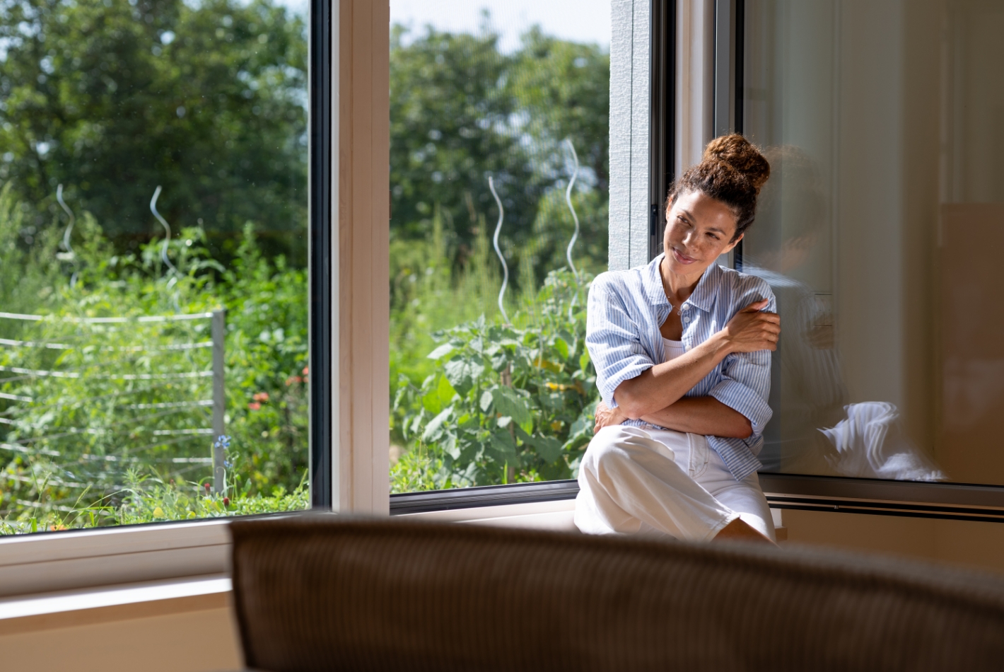 Frau sitzt entspannt auf einer Fensterbank vor einem großen, geöffneten Fenster mit grünem Garten im Hintergrund. Ein feinmaschiges Insektenschutz‑Gewebe ist im Fensterrahmen quasi unsichtbar und lässt viel Tageslicht und frische Luft hinein.