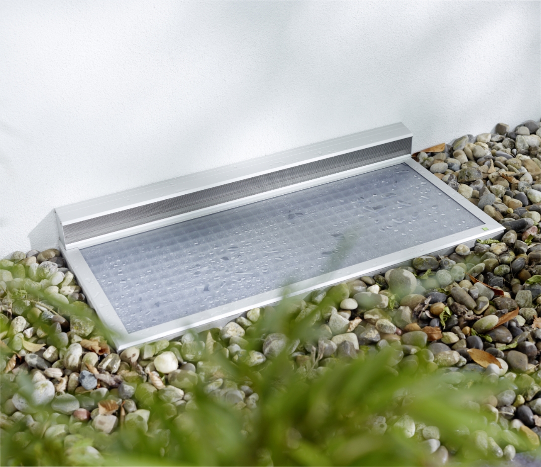 RESi Regenschachtabdeckung mit Belüftungselement an der Fassade. Transparente, wasserabweisende Fläche mit Tropfen, in Kiesbett eingebettet.