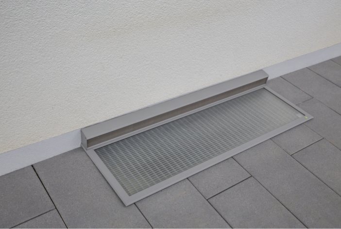 RESi Regenschachtabdeckung mit Belüftungskanal an der Hauswand, flach und bündig in graue Terrassenplatten integriert.