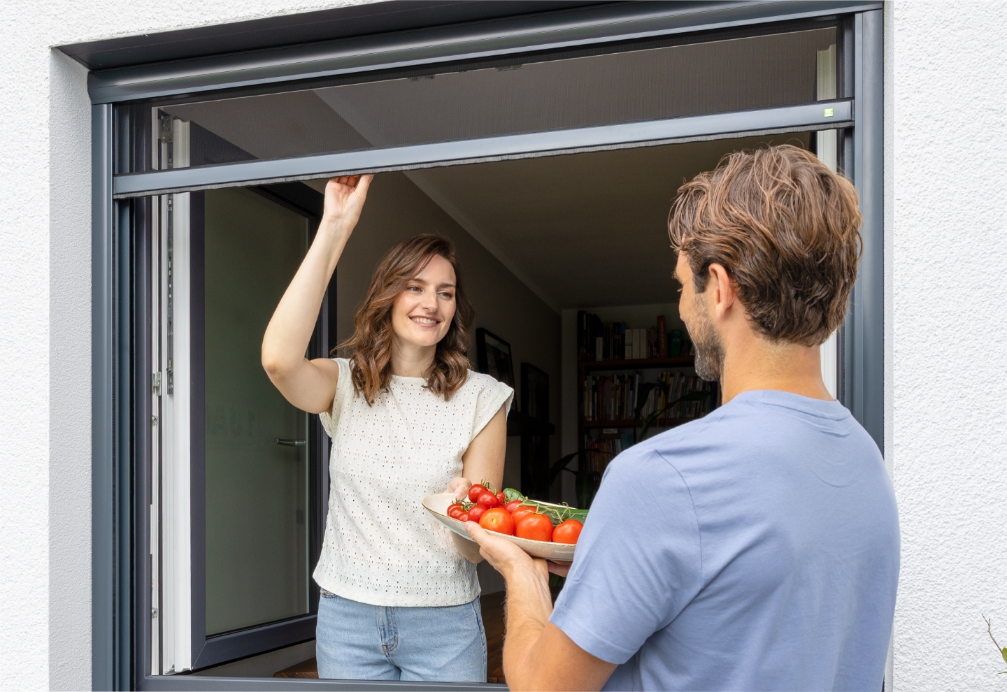 Eine Frau bedient ein Insektenschutz-Rollo an einem Fenster von innen, während ein Mann ihr einen Teller mit frischen Tomaten vom Garten aus reicht.