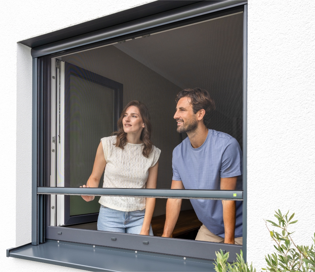 Zwei Personen stehen an einem geöffneten Fenster mit integriertem Insektenschutz-Rollo und schauen nach draußen.