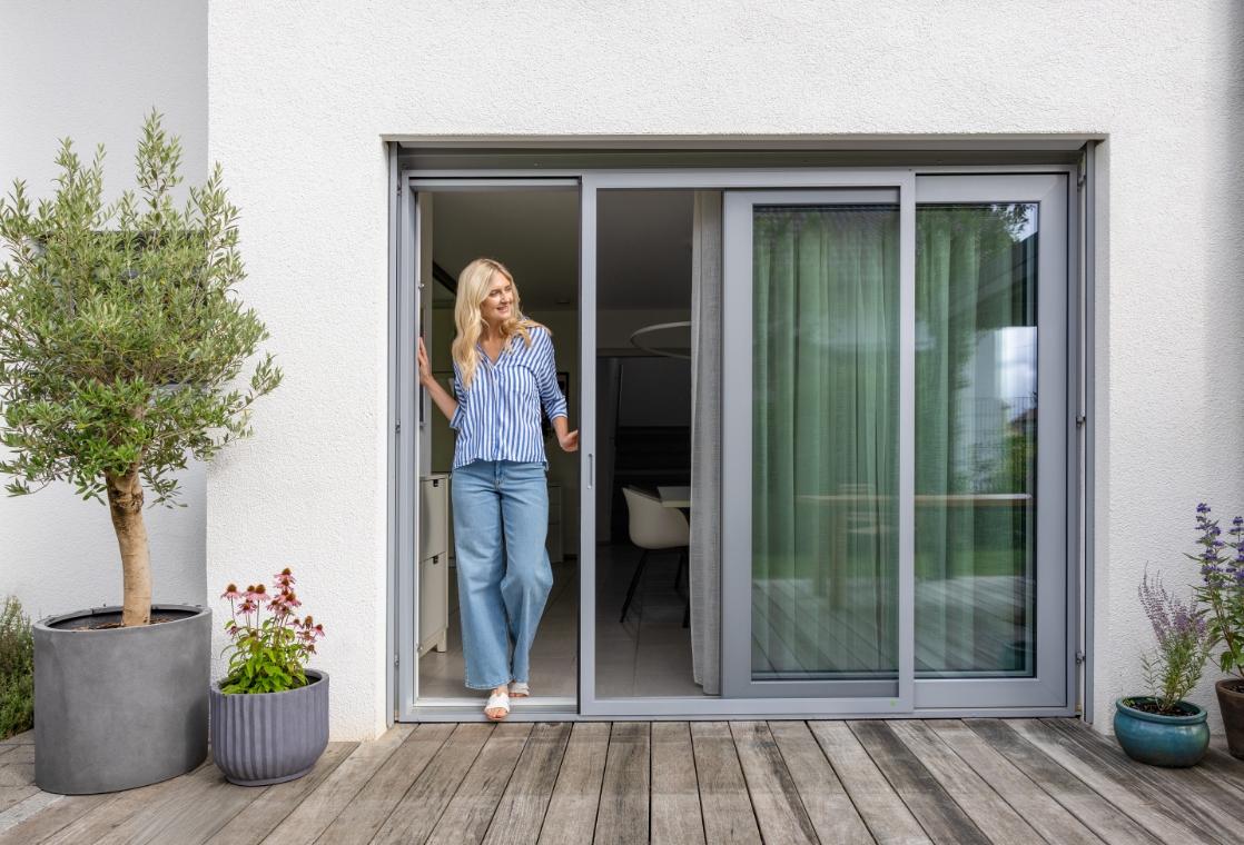 Frau öffnet eine Terrassentür mit Neher Insektenschutz Schiebeanlage; schlanker Rahmen, große Glasflächen, barrierefreier Zugang zur Terrasse, zuverlässiger Schutz vor Insekten.