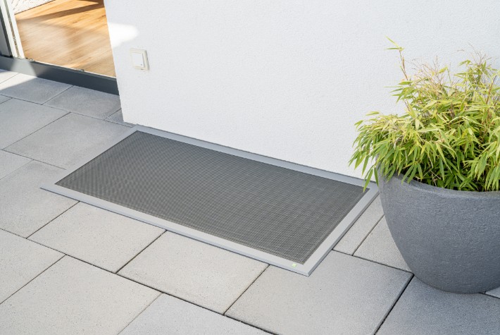 Neher Lichtschachtabdeckung auf moderner Terrassenfläche; flache, begehbare Abdeckung mit stabilem Rahmen und feinmaschigem Gitter, bündig am Hausanschluss montiert.