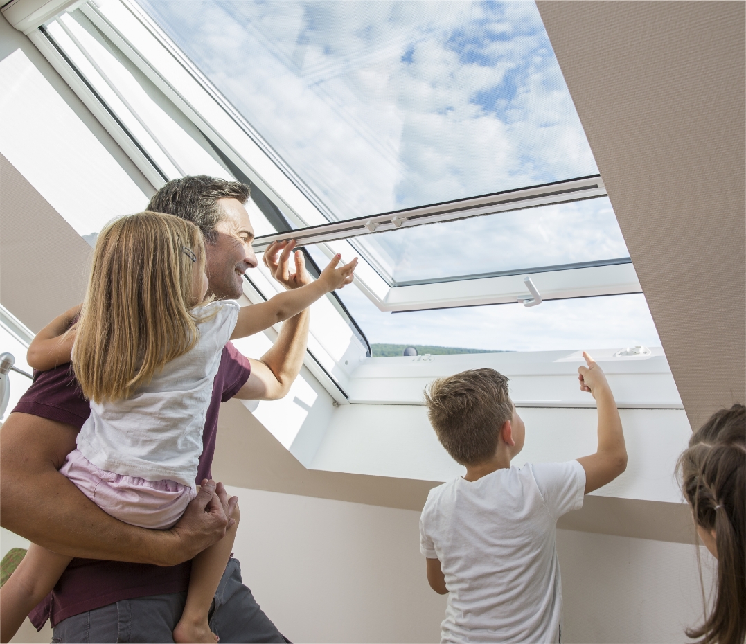Mann hält ein Kind auf dem Arm und öffnet mit der anderen Hand ein Fliegengitter-Rollo für das Dachfenster. Ein weiteres Kind streckt die Hand aus dem Fenster, damit ein Marienkäfer hinausfliegen kann.