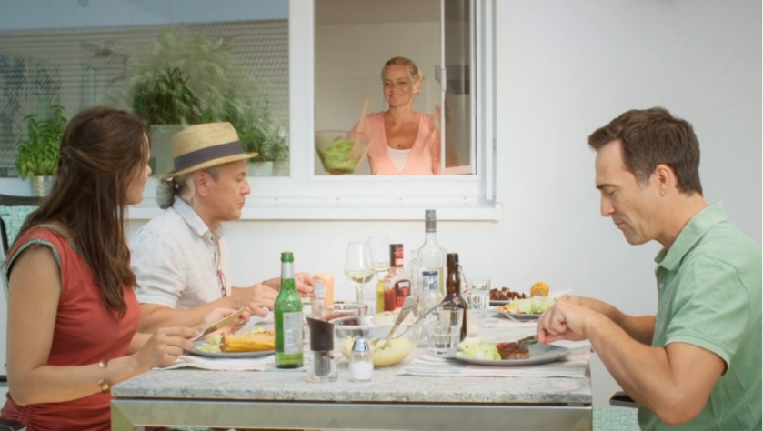 Familie sitzt beim Essen im Garten, während eine Person durch ein Pendelfenster mit Insektenschutz Speisen reicht.